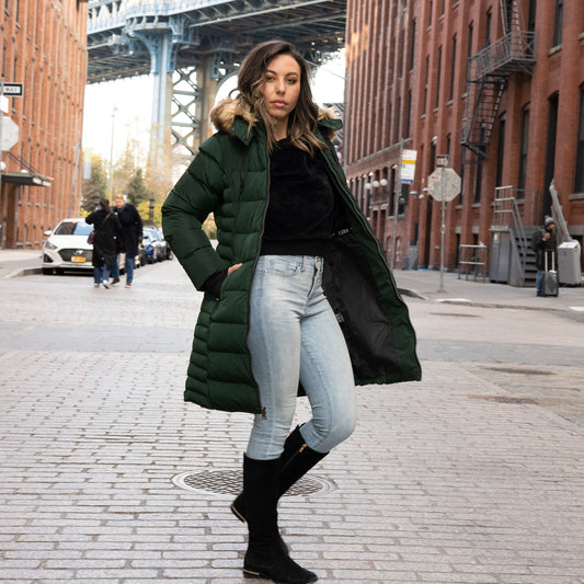 Woman in a stylish green mid-length winter parka with a faux fur hood, standing on a cobblestone street in DUMBO, Brooklyn.