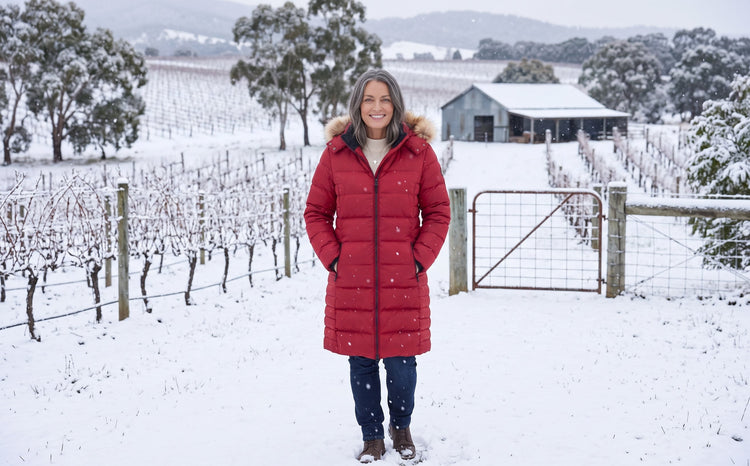 Woman in red winter coat with hands in pockets standing in a snow-covered vineyard.