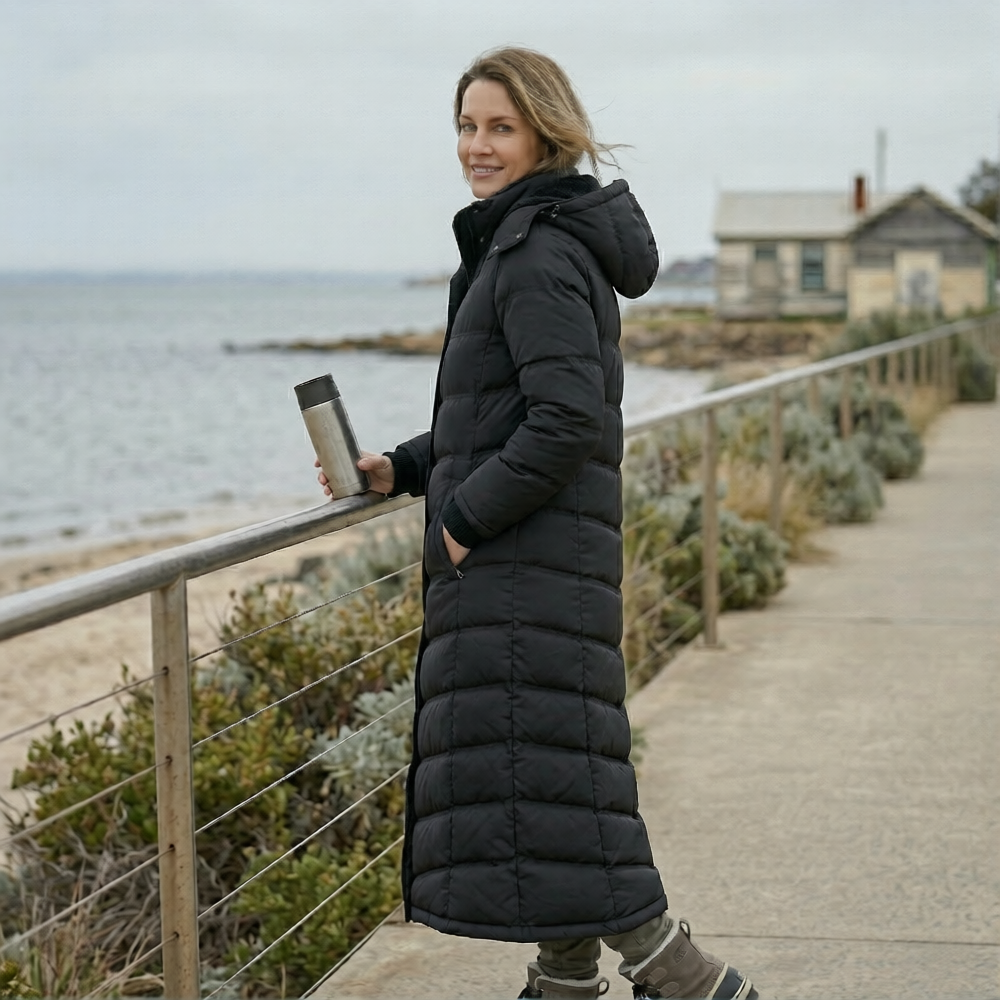 Woman in a long black puffer coat holding a thermos by the beach.