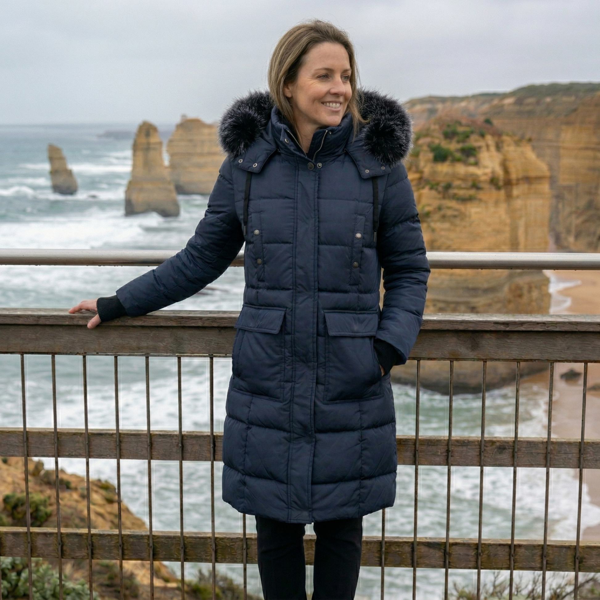 Woman in a blue coat with fur hood standing at a scenic location with ocean and cliffs.