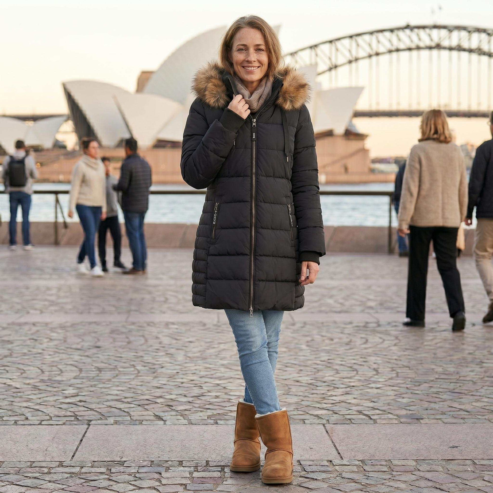 A woman in a black puffer coat and tan boots standing in front of the Sydney Opera House. She is wearing blue jeans and a scarf, with her right hand near her collar. The Opera House sails and the Harbour Bridge are in the background under a warm, late afternoon sky, with blurred other pedestrians.