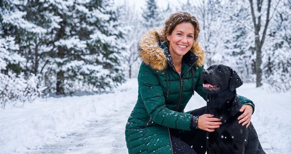 Mid Length Womens Winter Coat | ELORA Technical Fleece-Lined Parka A woman in a dark green winter coat with a brown fur-trimmed hood crouches in a snowy outdoor setting next to a large black dog.