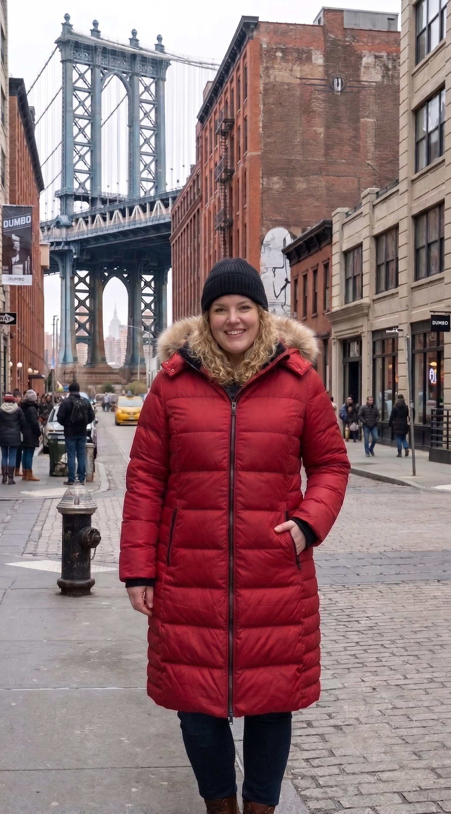 A woman wearing a long red puffer coat with a faux-fur trimmed hood and a black beanie stands on a city sidewalk with the Manhattan Bridge in the background.Women Plus Size Winter Coat with Hood