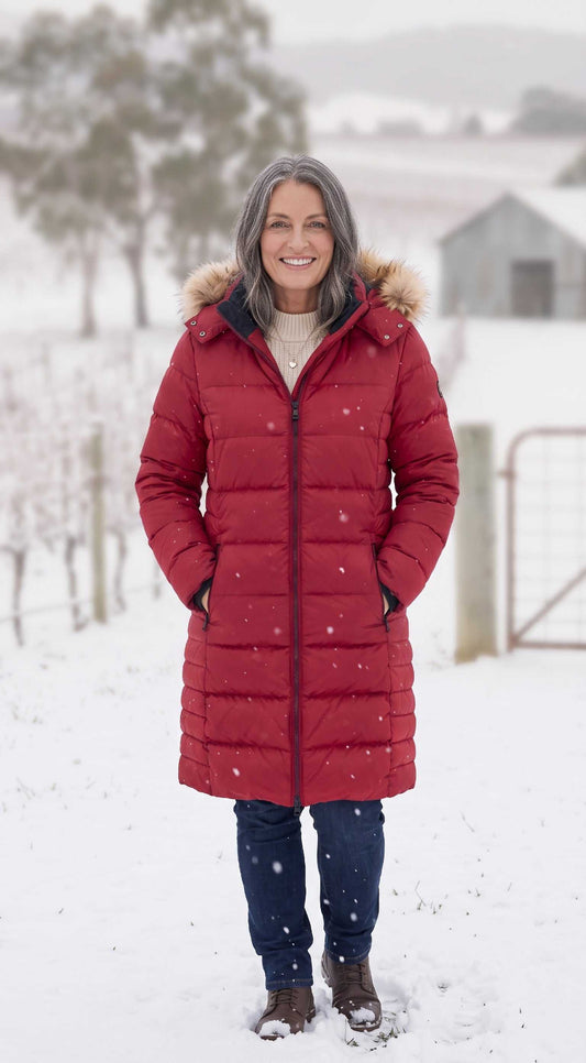 A full-body photograph of a woman with grey hair standing in a snow-covered vineyard. She is wearing a long, red puffer coat with a fur-trimmed hood, a white turtleneck sweater, blue jeans, and brown leather boots. She is smiling directly at the camera with her hands in her pockets. The background shows snow-dusted grapevines, trees, and a barn.