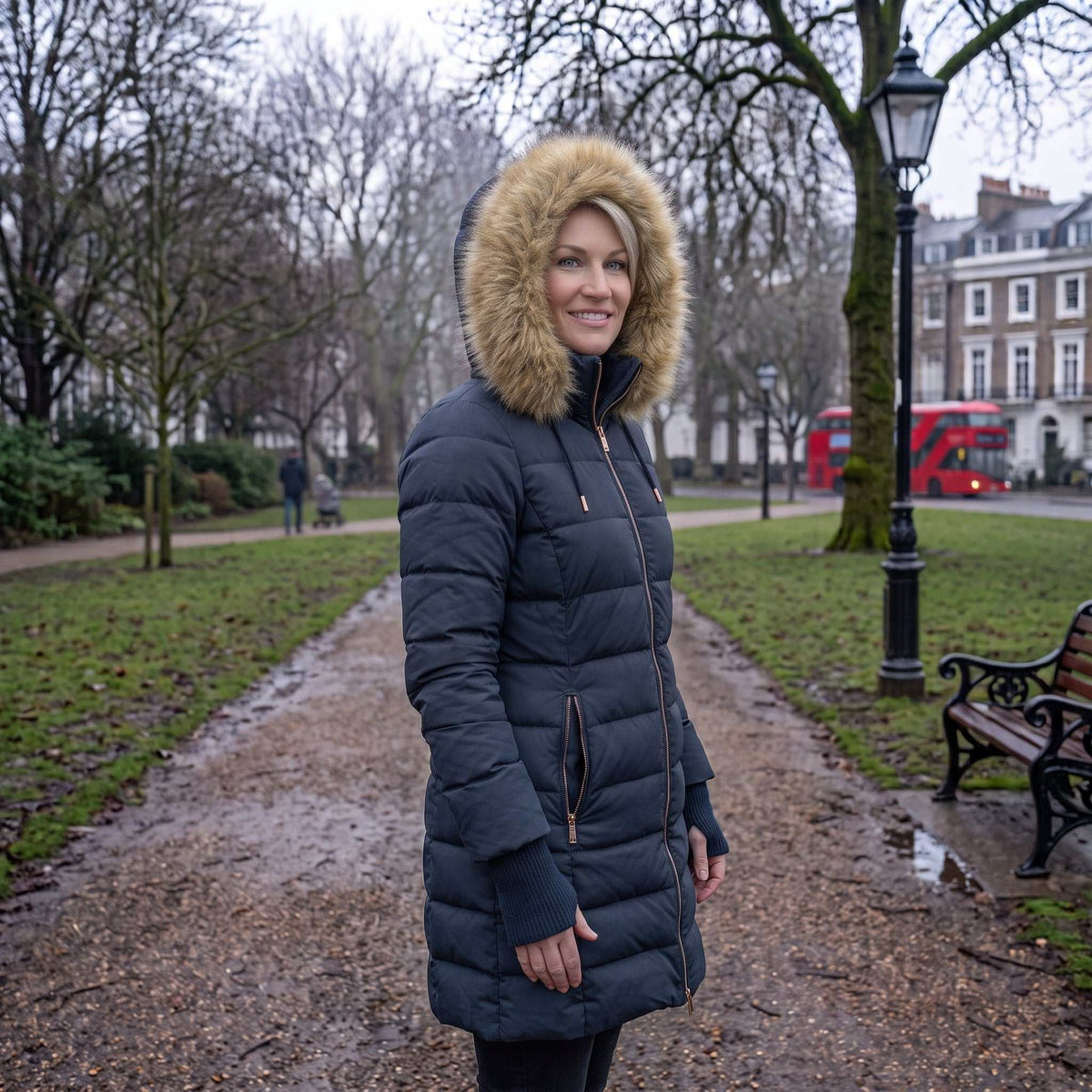 A woman wearing a Mid Length Womens Winter Coat | ELORA Technical Fleece-Lined Parka with a large tan faux-fur trimmed hood stands on a muddy park path. Bare trees, grass, benches, and a red double-decker bus are visible in the background.