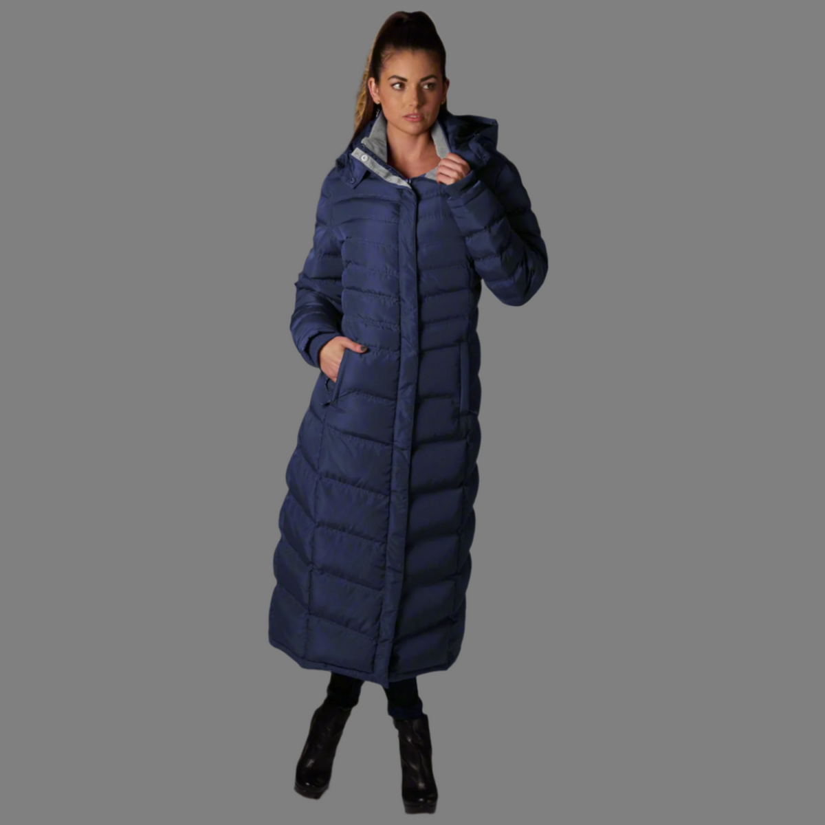 Women's navy blue full-length puffer coat with horizontal quilting, hood, zippered front, and hand pockets worn by model