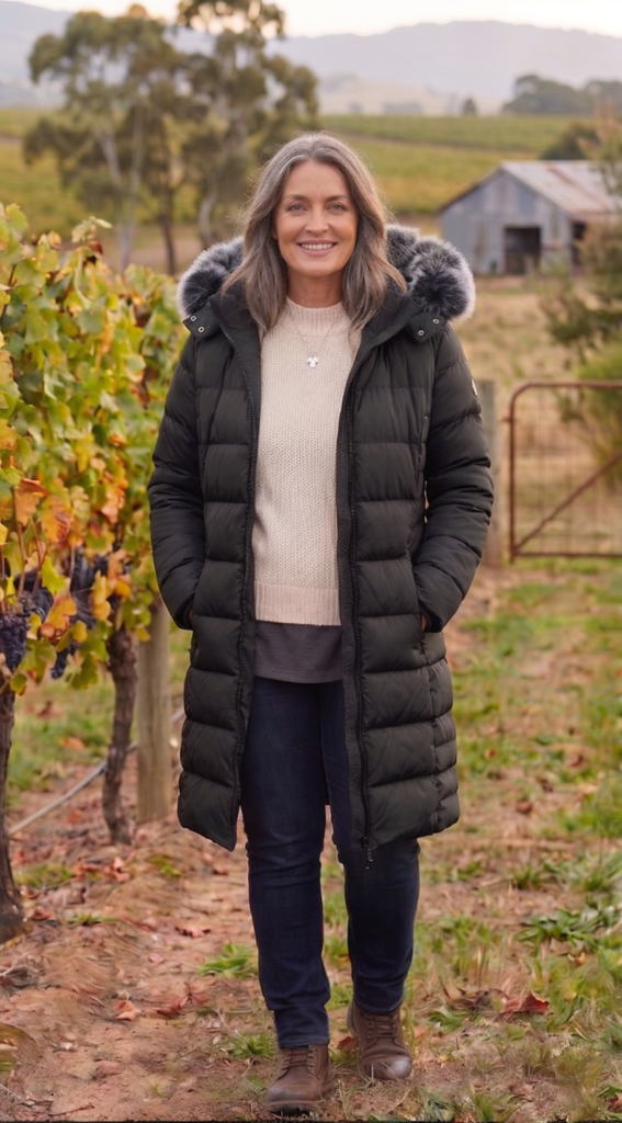 A woman with long grey hair smiling, standing in a vineyard wearing a long black quilted puffer coat with a salt-and-pepper faux fur-trimmed hood. Her hands are tucked into the coat pockets, and she is wearing dark jeans and brown leather boots. The background features grapevines, a rustic shed, and rolling hills under an overcast sky